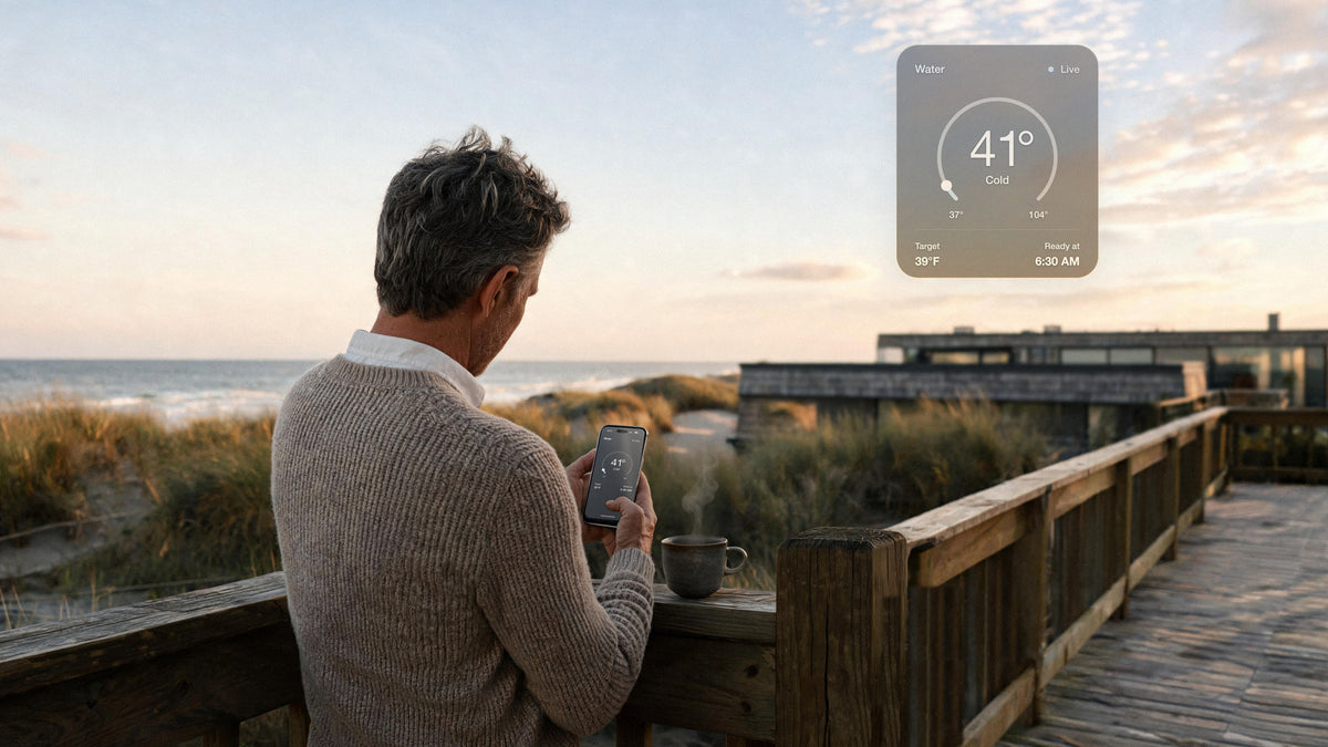 A man with salt-and-pepper hair in an oatmeal cashmere sweater checks his phone at the wooden rail of his beach house at dawn, a steaming stoneware cup beside him; a floating panel shows the water at 41° and ready at 6:30 AM.