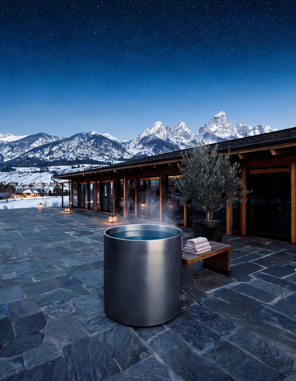 AURUM cylindrical vessel in Satin Steel set warm on a slate terrace at night, the Tetons beyond, steam rising