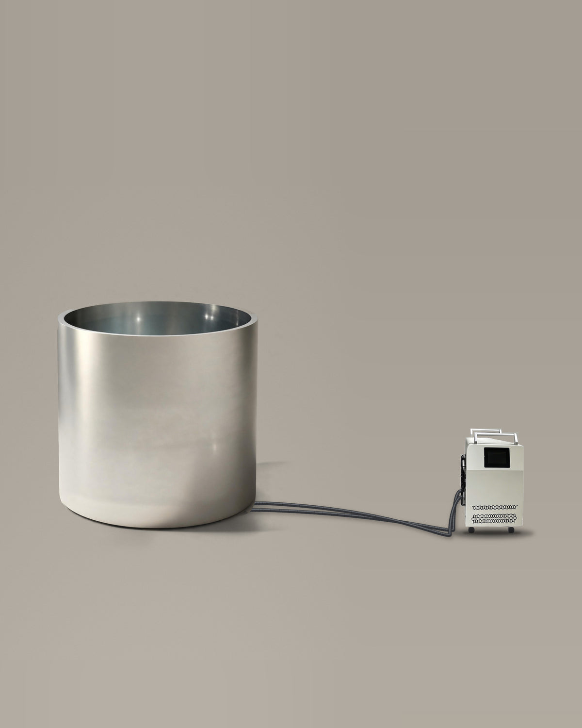 Brushed stainless steel cylindrical bathing vessel on a warm grey studio backdrop beside its Hot & Cold Unit, connected by two braided hoses