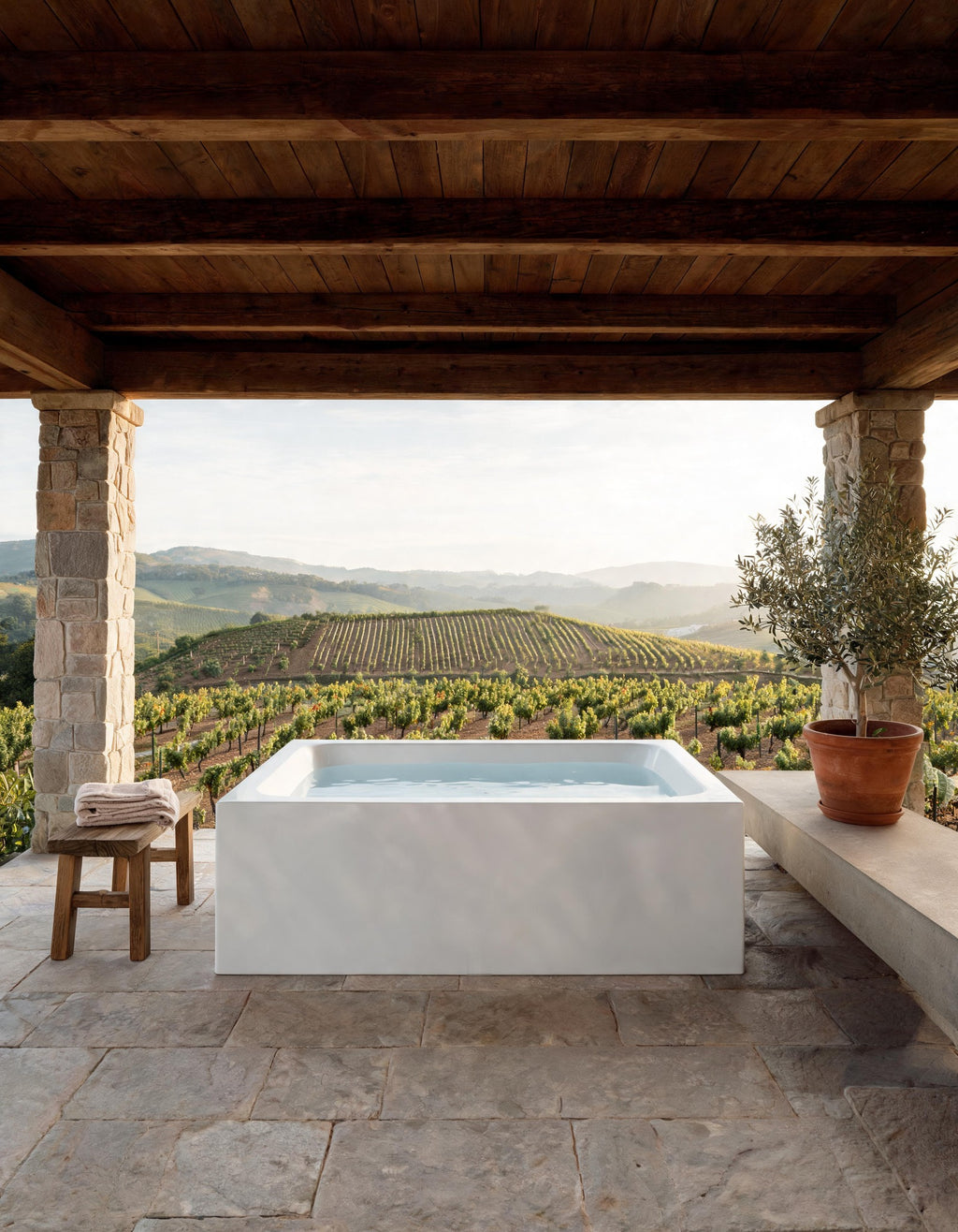 White rectangular bathing vessel on a covered stone terrace overlooking vineyards, an olive tree and a wooden stool beside it