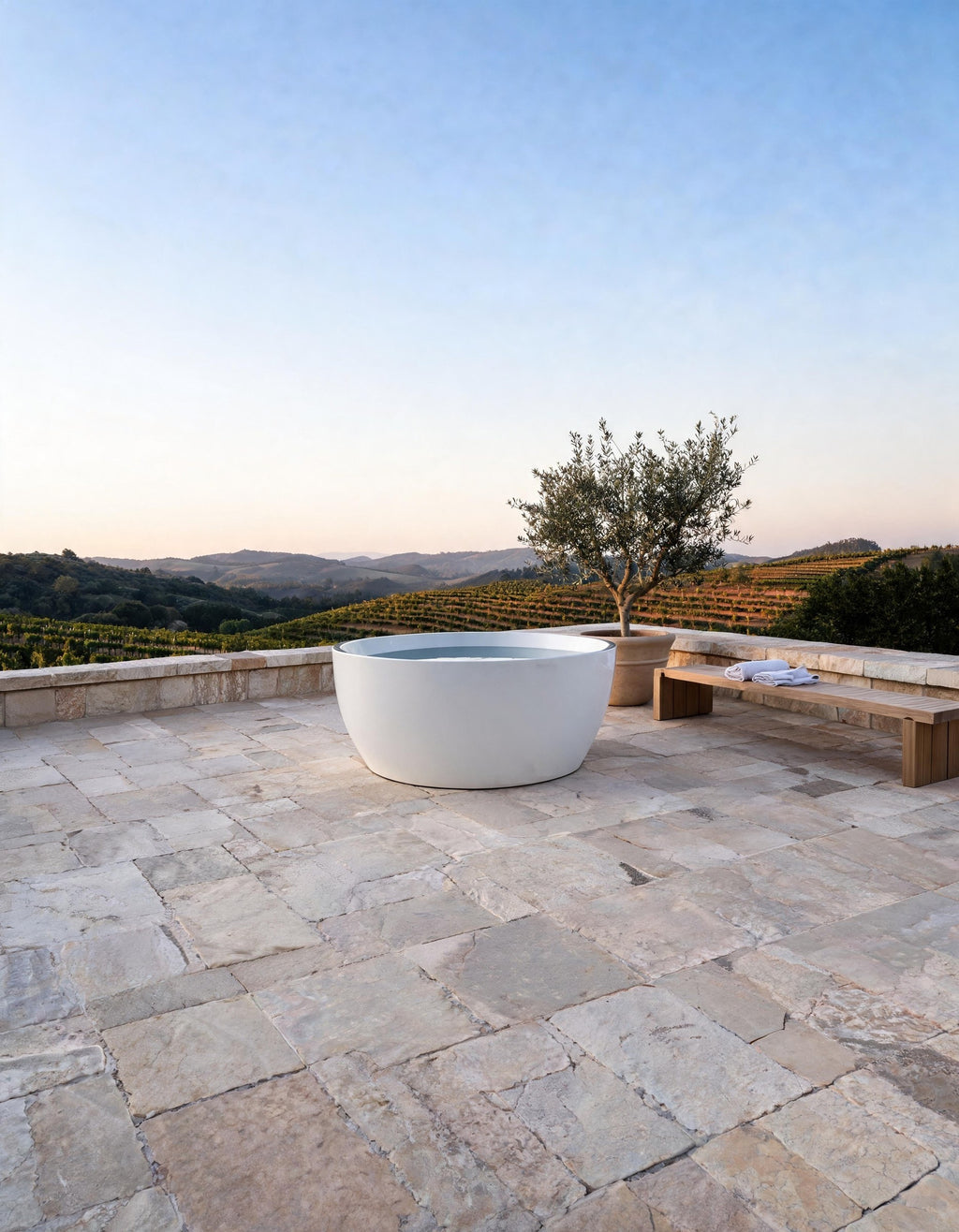 LUNA round vessel in white acrylic filled with cold water on an open stone terrace among vines, a bench and an olive tree beside it
