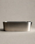 OBLONG hot and cold vessel, a long oval in Satin Steel, side view on a warm grey studio background